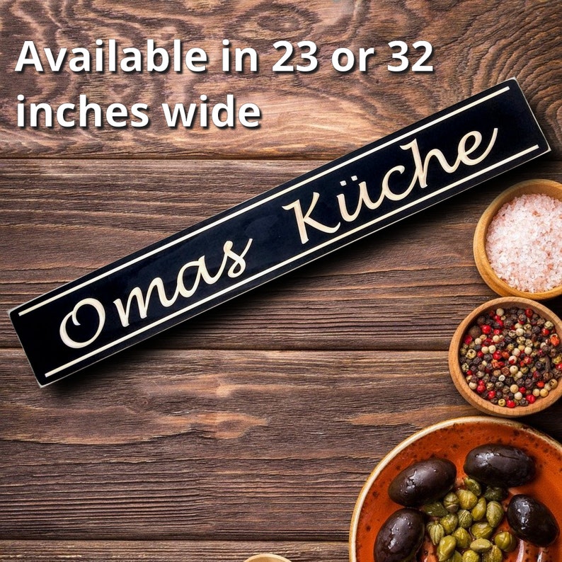 May include: A black wooden sign with gold lettering that reads "Omas K&uuml;che" on a rustic wooden background. The sign is available in 23 or 32 inches wide.