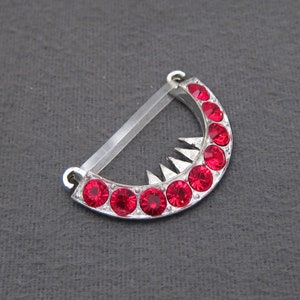 May include: A silver crescent-shaped pendant with a row of red rhinestones. The pendant has a pointed edge and a clear plastic loop for attaching to a chain.