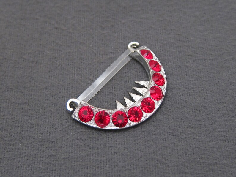 May include: A silver crescent-shaped pendant with a row of red rhinestones. The pendant has a pointed edge and a clear plastic loop for attaching to a chain.