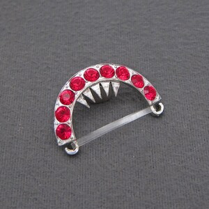 May include: A silver-toned, crescent-shaped brooch with a row of red rhinestones. The brooch has a pointed design in the center and a clear plastic clasp.