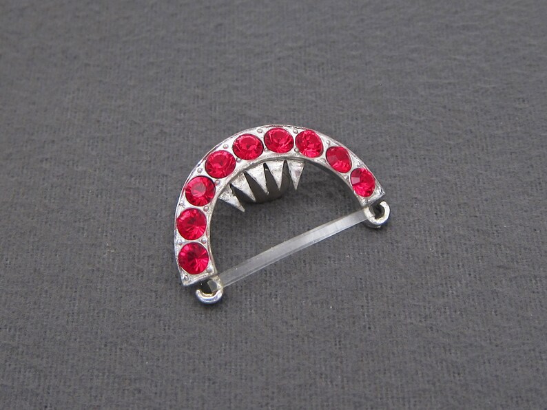 May include: A silver-toned, crescent-shaped brooch with a row of red rhinestones. The brooch has a pointed design in the center and a clear plastic clasp.