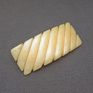 May include: A light-colored, rectangular hair clip with a segmented design. The clip features a series of angled, light-colored segments held together by thin, gold-colored metal accents. The overall shape is slightly curved, and the clip appears to be made of wood or a similar material.
