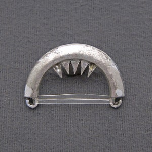 May include: A silver-toned metal buckle with a curved shape and a row of triangular teeth. The buckle has two loops on either side.