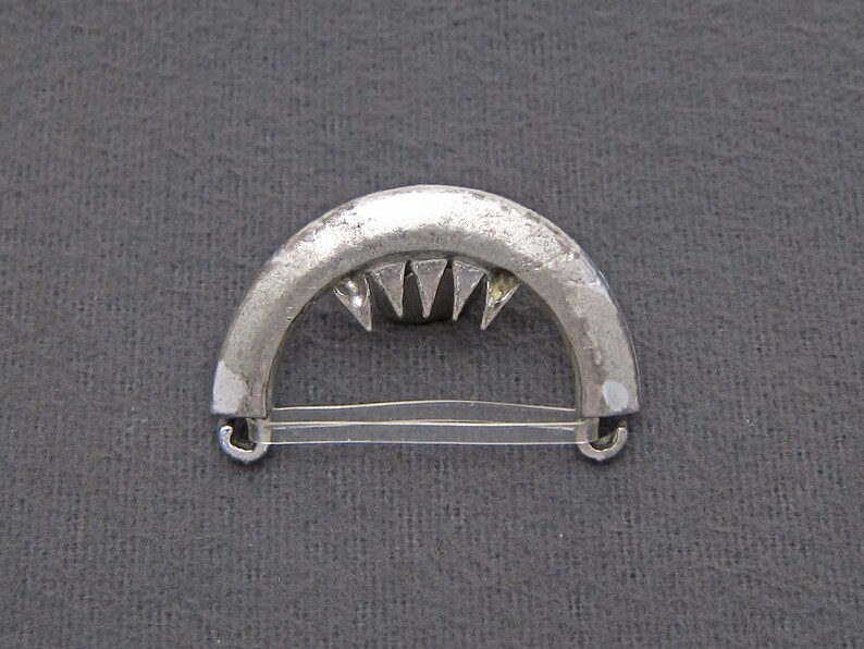 May include: A silver-toned metal buckle with a curved shape and a row of triangular teeth. The buckle has two loops on either side.