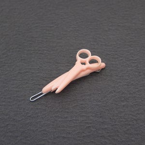 May include: A pair of light pink hair styling scissors with a silver metal clip. The scissors have two finger holes and a curved blade. The scissors are laying on a dark gray surface.