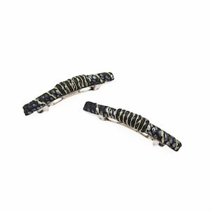 May include: Two black and gold barrettes with a textured pattern. The barrettes have a curved shape and are decorated with gold wire. The background is white.