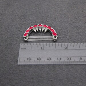 May include: A silver hair clip with a row of red rhinestones in the shape of an arch. The clip is about 1 inch wide and has a clear elastic band.