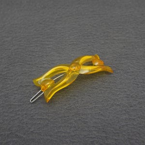 May include: A translucent yellow hair clip with a curved design. The clip features a silver metal clasp and is set against a gray background. The hair accessory is a decorative item.
