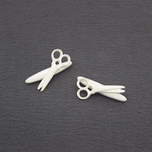 May include: Two ivory-colored scissors are displayed against a dark gray background. The scissors have a classic design with rounded finger holes and pointed blades. The blades are closed, and the scissors are positioned at different angles.