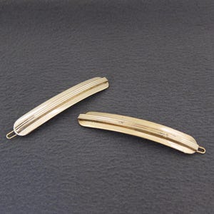 May include: Two gold-colored barrettes with a curved design and horizontal lines. The barrettes have a simple, elegant style and are set against a dark gray background. One is slightly larger than the other.