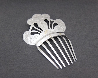 May include: A silver-toned hair comb with a decorative floral design. The comb features a fan-shaped top with cut-out details and engraved floral patterns. The comb's teeth are straight and evenly spaced.