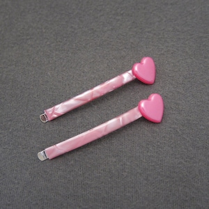 May include: Two pink hair clips with heart-shaped embellishments. The clips are made of a marbled plastic material.
