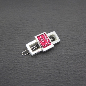 May include: A silver-toned metal barrette with a rectangular, magenta-colored center piece. The center piece has white text. The barrette is composed of two rectangular frames. The barrette is on a dark gray surface.