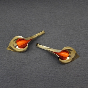 May include: Two gold-colored, leaf-shaped earrings with an orange, teardrop-shaped center. The earrings are set against a dark gray background, creating a striking contrast. The design is elegant and modern.