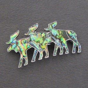 May include: A silver brooch featuring three moose silhouettes crafted from iridescent abalone shell. The moose are positioned in a line, with their heads facing the same direction. The brooch exhibits a green, blue, and purple iridescent sheen.
