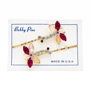 May include: Two gold-coloured bobby pins with dragonfly designs. Each dragonfly has red, pink, and light blue rhinestones. The bobby pins are on a white card with the text "Bobby Pins" and "MADE IN U.S.A."