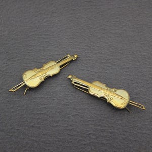 May include: Two gold-colored violin-shaped hair clips. The clips are detailed with the shape of a violin, including the strings and scroll. The background is a dark gray, textured surface.