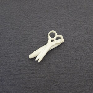 May include: Ivory-colored scissors with a classic design, featuring two finger holes and a pointed blade. The scissors are made of a solid material and rest on a dark gray surface. The overall aesthetic is simple and elegant.