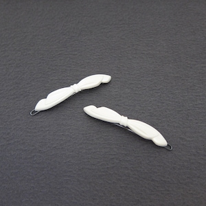 May include: Two white hair barrettes with a bow-like design. Each barrette has a curved shape with a small metal clasp. The barrettes are lying on a dark gray surface.