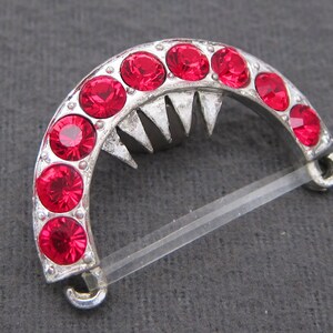 May include: A silver-toned metal clasp with a half-circle design. The clasp is adorned with a row of red rhinestones.