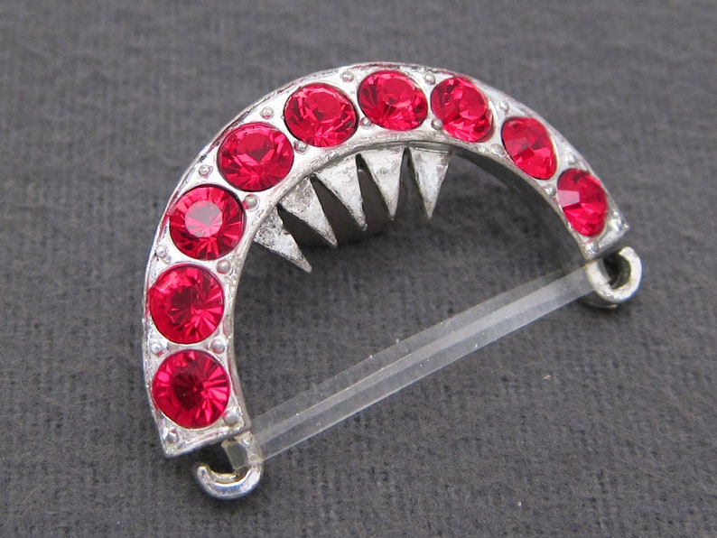 May include: A silver-toned metal clasp with a half-circle design. The clasp is adorned with a row of red rhinestones.