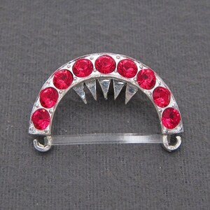 May include: A silver-toned hair comb with a half-circle design. The comb is decorated with red rhinestones and has a pointed edge.