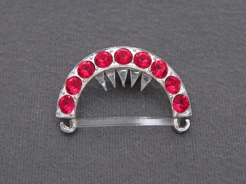 May include: A silver-toned hair comb with a half-circle design. The comb is decorated with red rhinestones and has a pointed edge.