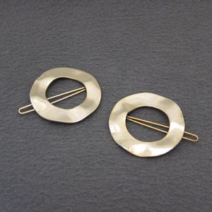 May include: Two gold-colored hair barrettes. Each barrette features a hammered, circular design with a gold-colored pin. The barrettes are displayed on a dark gray surface.