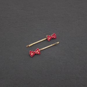 May include: Two gold-toned bobby pins, each adorned with a bow-shaped cluster of small, vibrant red rhinestones. The pins are set against a dark gray background, creating a striking contrast.