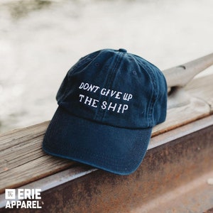 May include: Navy blue baseball cap with the embroidered text "DON'T GIVE UP THE SHIP" in white. The cap is sitting on a wooden surface with a blurred water background. The Erie Apparel logo is visible.