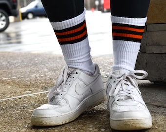May include: A pair of white crew socks featuring two horizontal orange stripes and one black stripe at the top, worn with white lace-up athletic sneakers. The socks are ribbed and extend to the mid-calf, paired with white leather shoes.