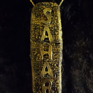 May include: A gold necklace with a pendant that says "SAHAR" in a stylized font. The pendant is made to look like an ancient Egyptian artifact.