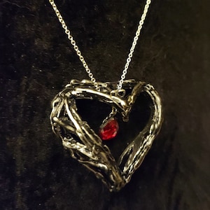 May include: A silver heart-shaped pendant necklace with a red gemstone hanging from the center. The pendant is made of intricate, twisted metal.