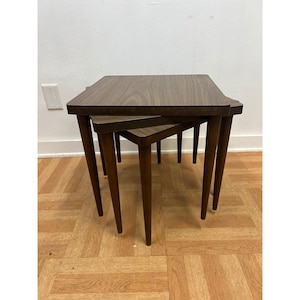 May include: A set of three nested tables featuring a dark brown wood grain finish. Each table has a square top with rounded corners and four tapered legs that extend outward from the base, providing a stable structure for the stacked arrangement.