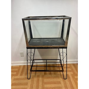 May include: A rectangular glass display case with a metallic frame, featuring a dark base and a black metal stand with four thin legs, decorative scrollwork accents on the sides, and a lower shelf constructed from a grid of thin metal bars.