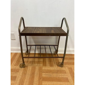 May include: A vintage serving cart with a dark wood-grain top and a lower shelf. The cart has a metal frame with curved handles and rolls on small wheels. The overall color scheme is brown and gold.
