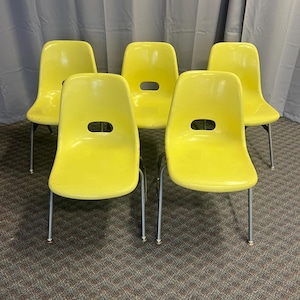 May include: Five yellow chairs with a curved back and a rounded opening near the top. The chairs have silver metal legs and are arranged in a group. The chairs are made of a glossy material.