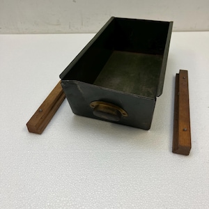 May include: A dark green metal drawer with a brass handle and two wooden supports. The drawer is rectangular and open, with a slightly distressed appearance. The wooden supports are rectangular and brown.