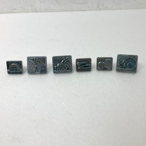 May include: A set of seven small, square, metal buttons with various embossed designs. The designs include a roller coaster, a mushroom, a frog, a pair of skis, a number 2, and a snail. The buttons are a muted gray color.