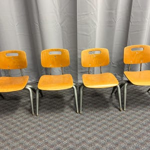 May include: Four vintage wooden chairs with light brown seats and backs. The backs have a rectangular cutout. The chairs have gray metal legs. The chairs are arranged in a row on a patterned carpet.