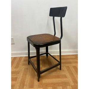 May include: An industrial-style chair with a black metal frame and a square seat. The seat has a brown, perforated surface. The chair has a small backrest and is designed with a minimalist aesthetic, suitable for a workshop or studio.