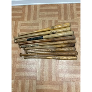 Puede incluir: Una colección de nueve bates de béisbol de madera dispuestos en formación paralela. Los bates presentan varios tonos de madera natural, desde el bronceado claro hasta el marrón oscuro, con varias marcas y textos de Louisville Slugger.