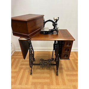 May include: A vintage treadle sewing machine featuring a black cast iron frame, a wooden tabletop with a drop-leaf side, and a matching wooden cover. The unit includes a set of three small drawers on the right side and a decorative metal foot pedal.
