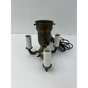 May include: A brass lamp base featuring a central cylindrical housing with three extending arms, each topped with a white socket. The fixture includes a black electrical cord and plug, with small screws visible on the central upper rim.