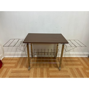 May include: A rectangular side table with a dark brown wood-grain top and a gold-toned metal frame. The table features two fold-out wire side shelves and a lower wire rack for storage, all supported by four small wheels for mobility.