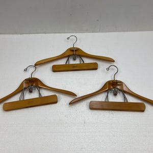 May include: Three wooden clothes hangers with metal hooks and clips. The hangers are a light brown color and have a classic design. The hooks are silver-colored. The hangers are designed for clothing.