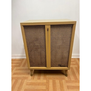May include: A rectangular wooden cabinet with a light tan finish, featuring two front panels covered in a textured brown woven fabric. A vertical wooden divider runs down the center, and the unit is supported by four tapered legs with metal tips.
