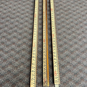 May include: Three vintage wooden measuring sticks, each marked with inch increments and numbered in red and black. The rulers are made of wood and are laid out on a textured gray surface.