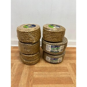 May include: Six coils of tan-colored poly rope are stacked on a wooden floor. Each coil is wrapped in clear plastic. The label on the side of the rope reads "POLY ROPE". The coils are tightly wound and appear durable.