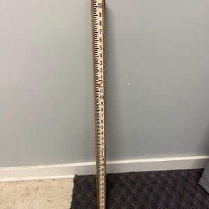 May include: A wooden measuring stick with black and red markings. The stick is marked from 1 to 11 inches, with each inch clearly labeled. The numbers are in red, and the lines are black. The stick is used for measuring.